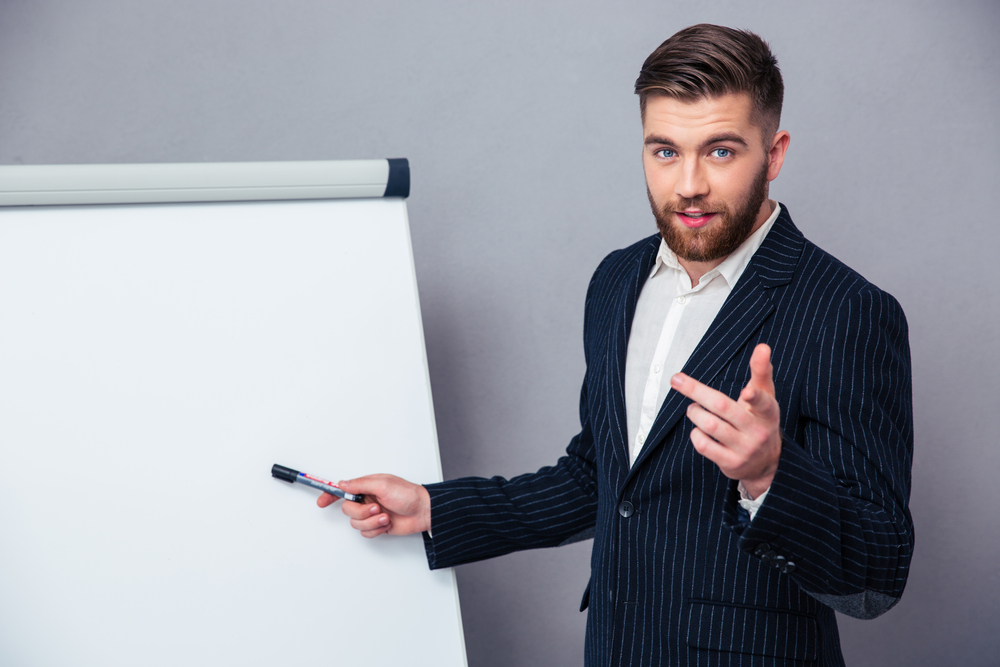 Portrait of a young businessman in suit presenting something on blank board over gray background